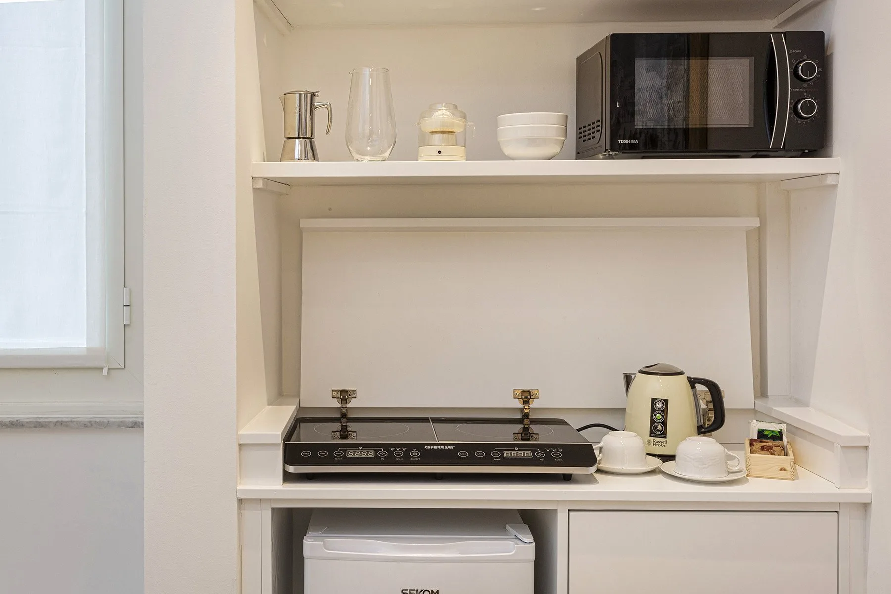 A compact kitchen area with a microwave, cooktop, and kettle on open shelves.