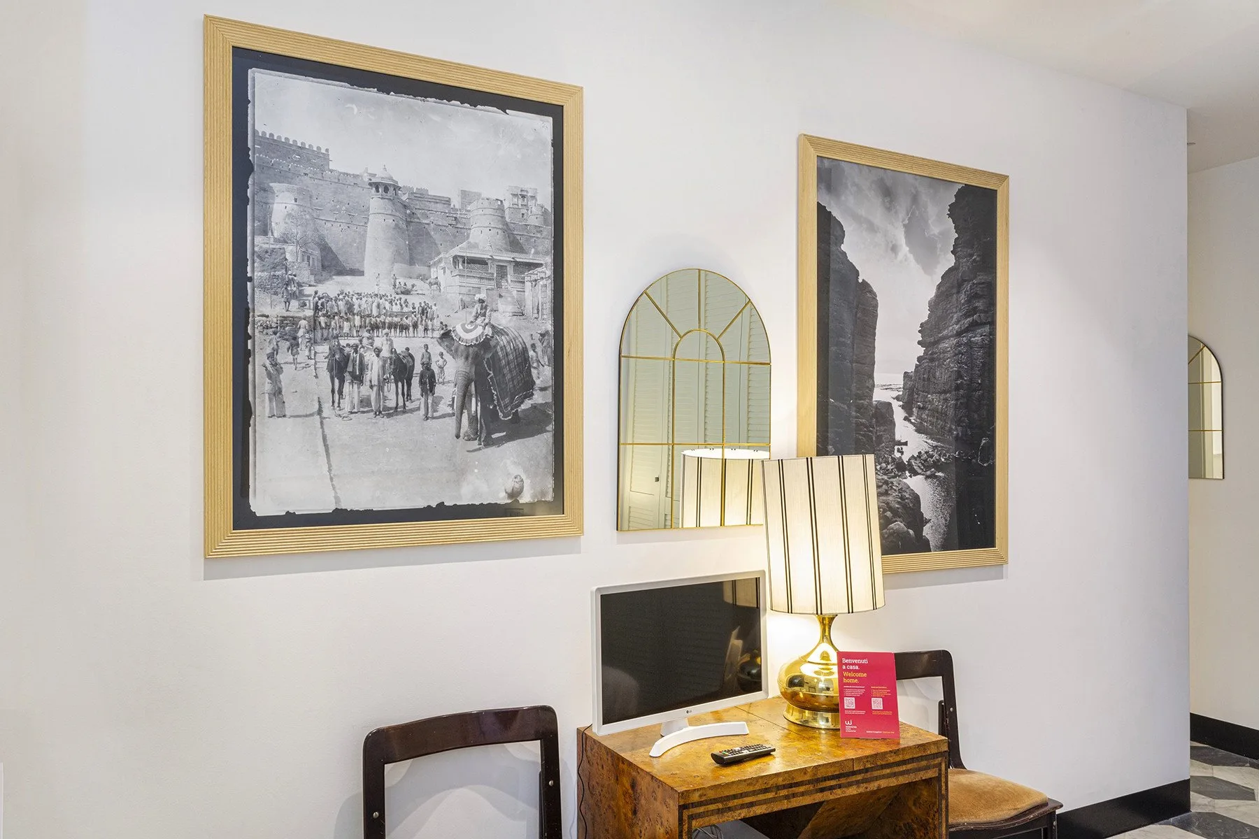 A room corner with framed black-and-white pictures above a wooden desk and lamp.