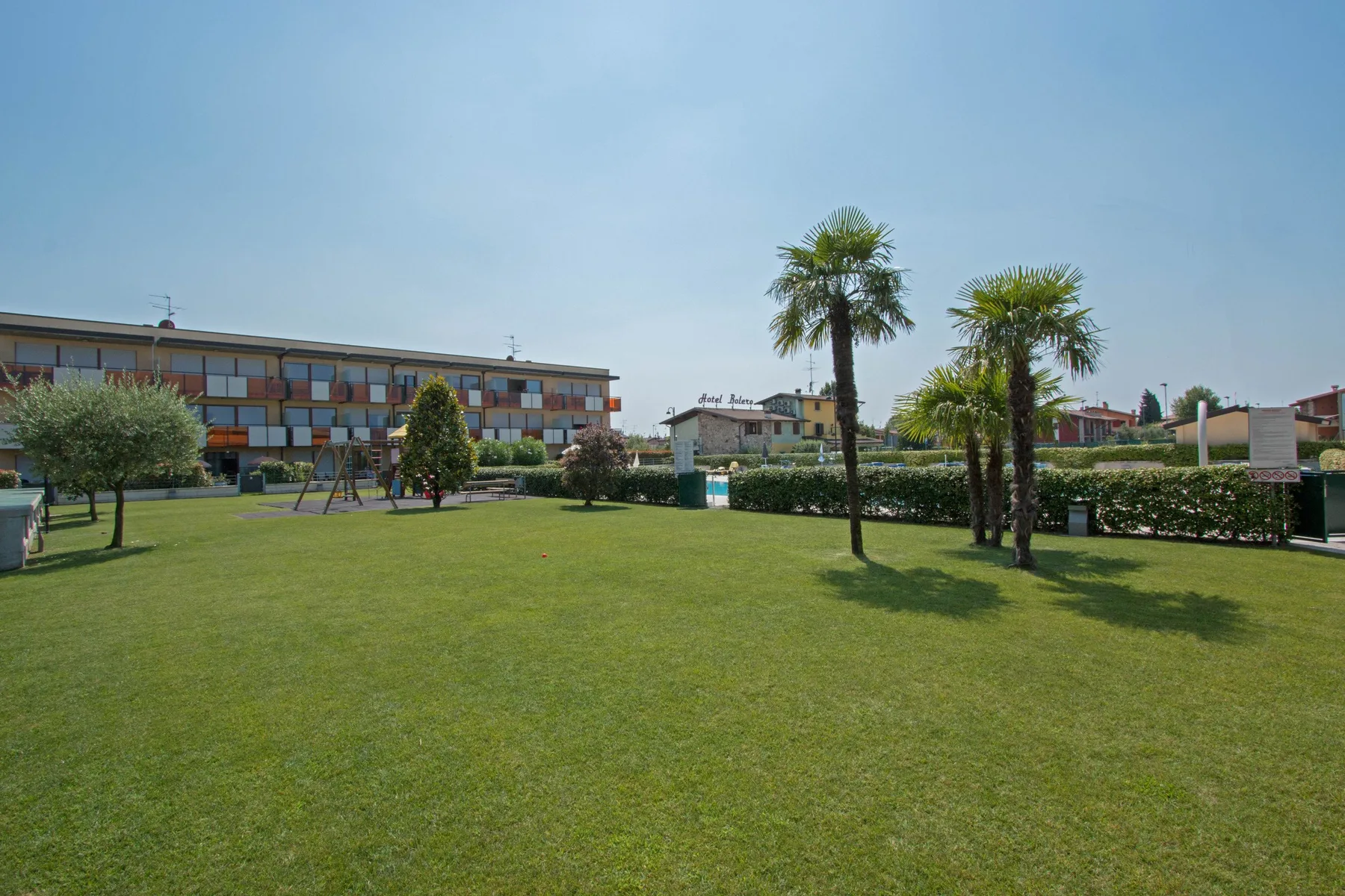 A grassy outdoor area with palm trees, shrubs, and a building in the background.