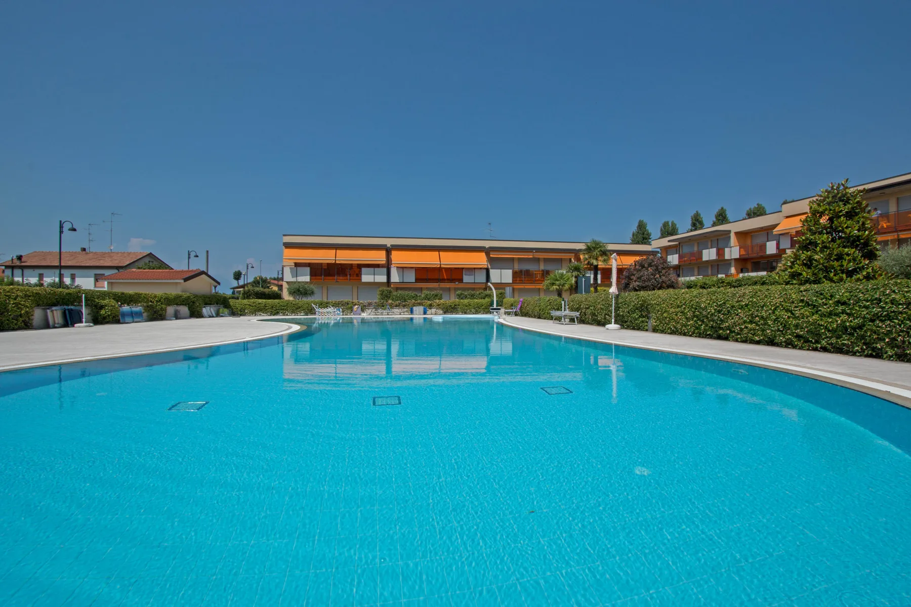 An outdoor swimming pool sits in front of low-rise buildings under a clear sky.