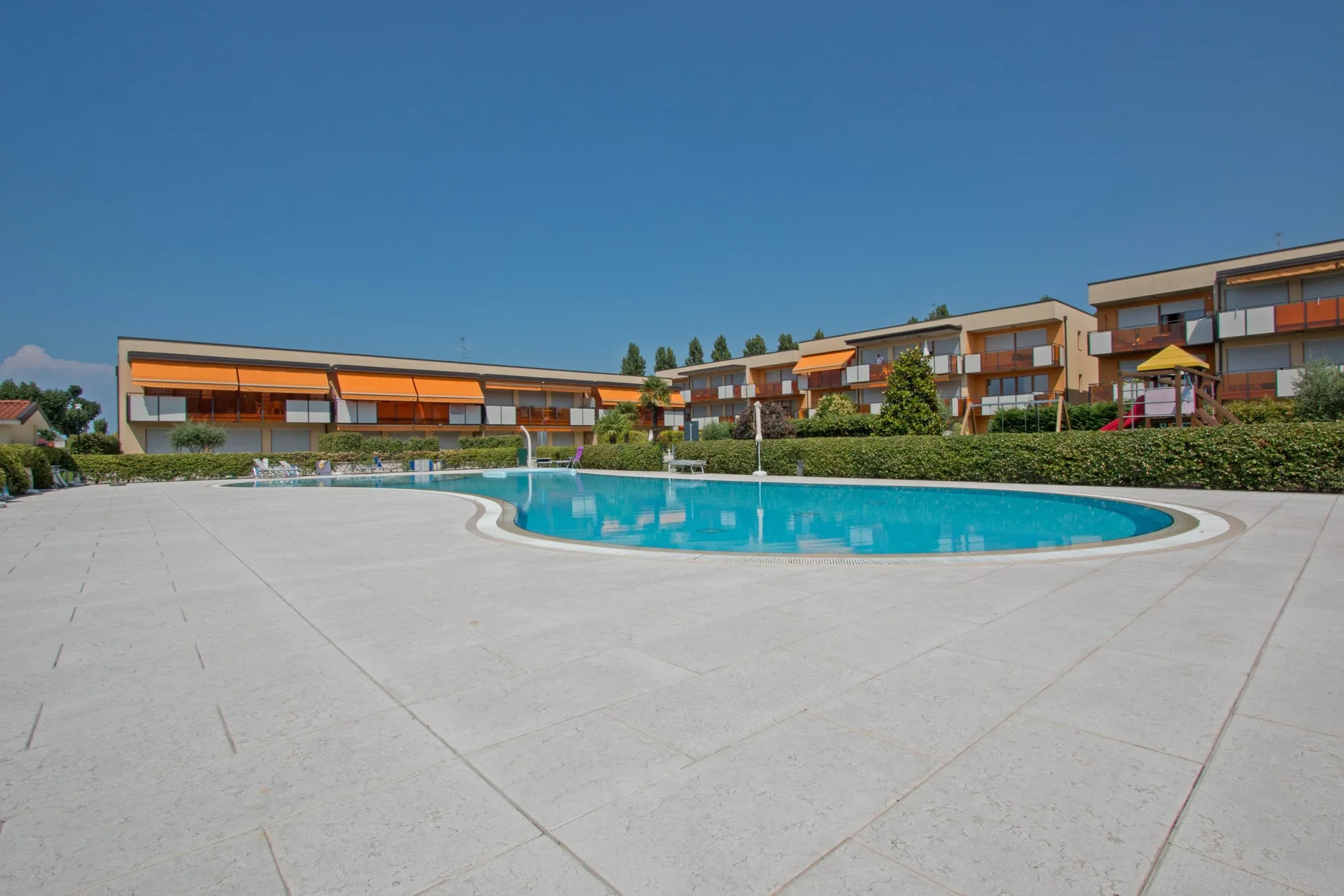 A swimming pool sits within a broad paved courtyard, with buildings and greenery beyond.