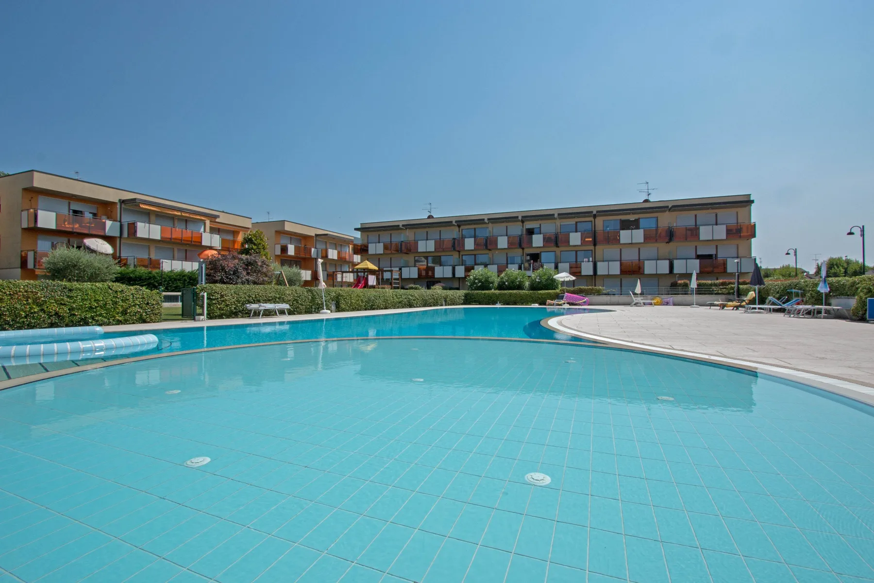 An outdoor swimming pool sits in front of low-rise buildings under a clear sky.