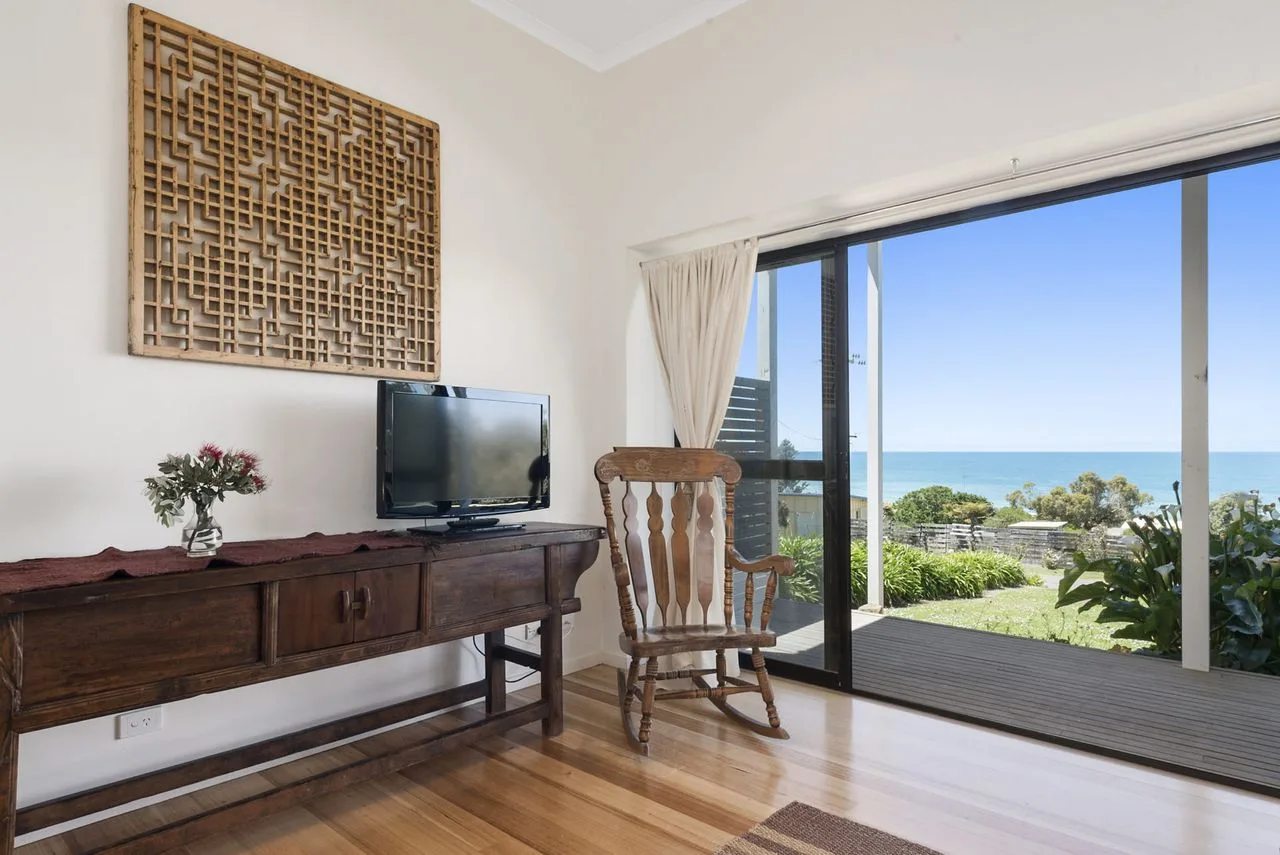 Una sala de estar con televisor, asiento de madera y puertas corredizas de vidrio con vista al océano.