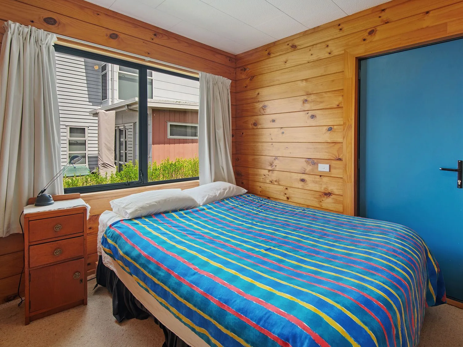 Un dormitorio con paredes de madera, una cama con ropa de cama azul a rayas, una mesita de noche con una lámpara y una ventana en Bay View Bach - Langs Beach Holiday Home, en Langs Beach
