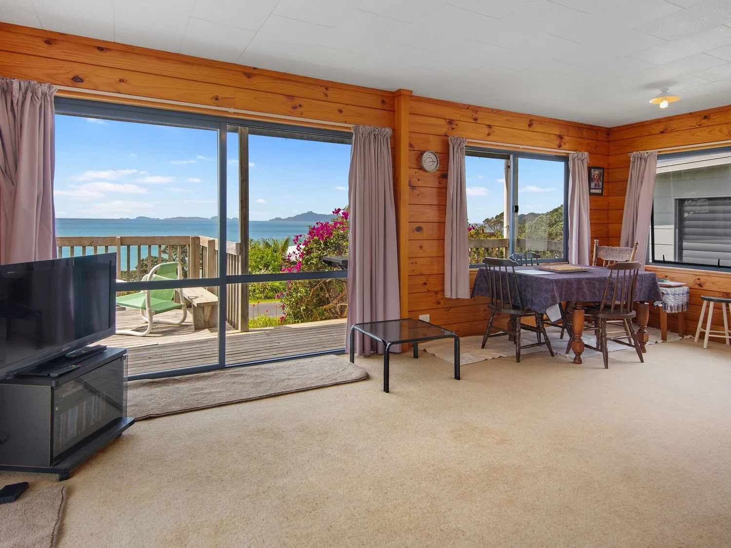 Una sala de estar con paredes de madera, una mesa de comedor con sillas, un televisor sobre un soporte y grandes ventanas con vistas al mar en Bay View Bach - Langs Beach Holiday Home, en Langs Beach