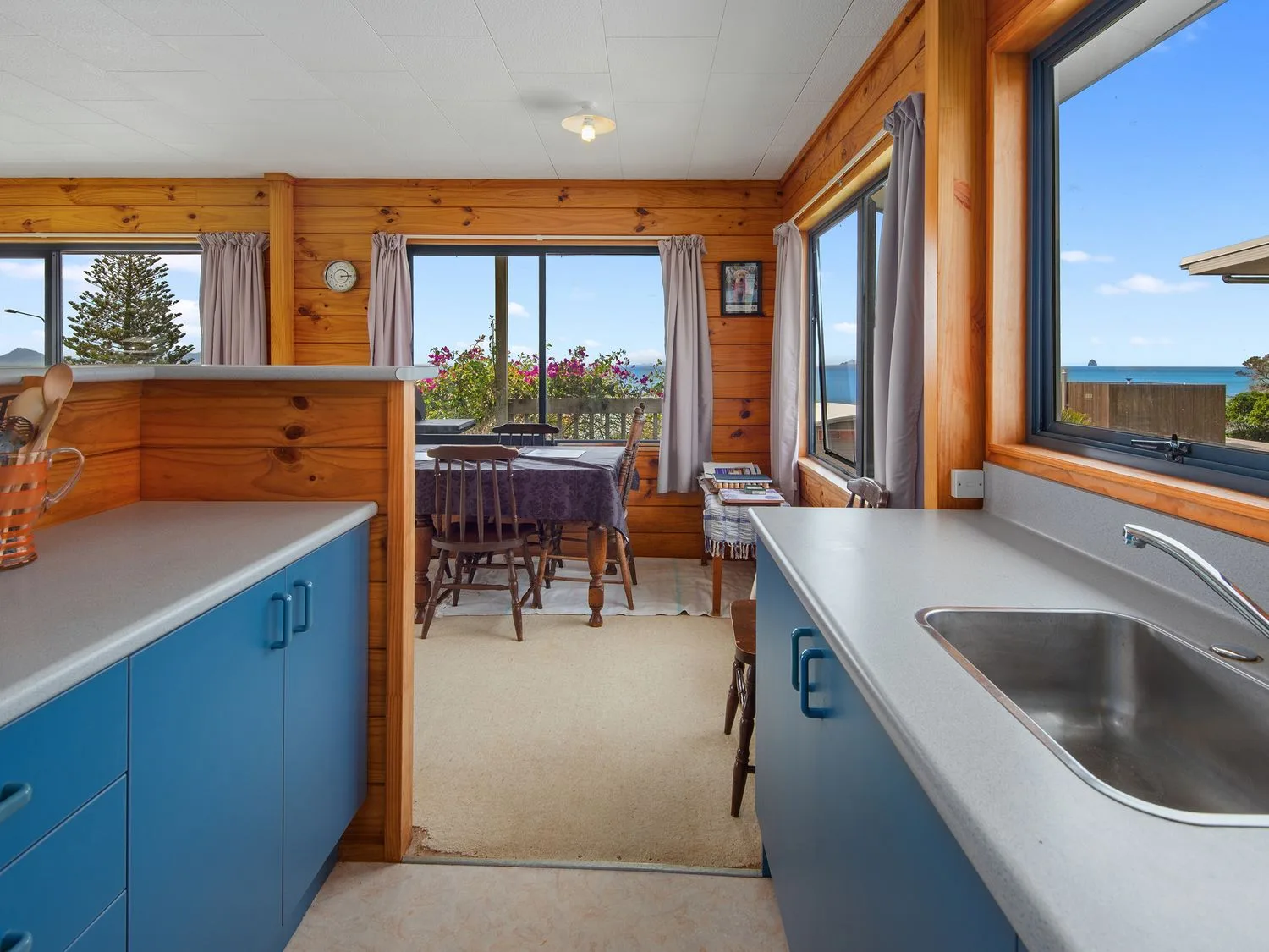 Una cocina con armarios azules y fregadero se abre a un comedor con una mesa.
