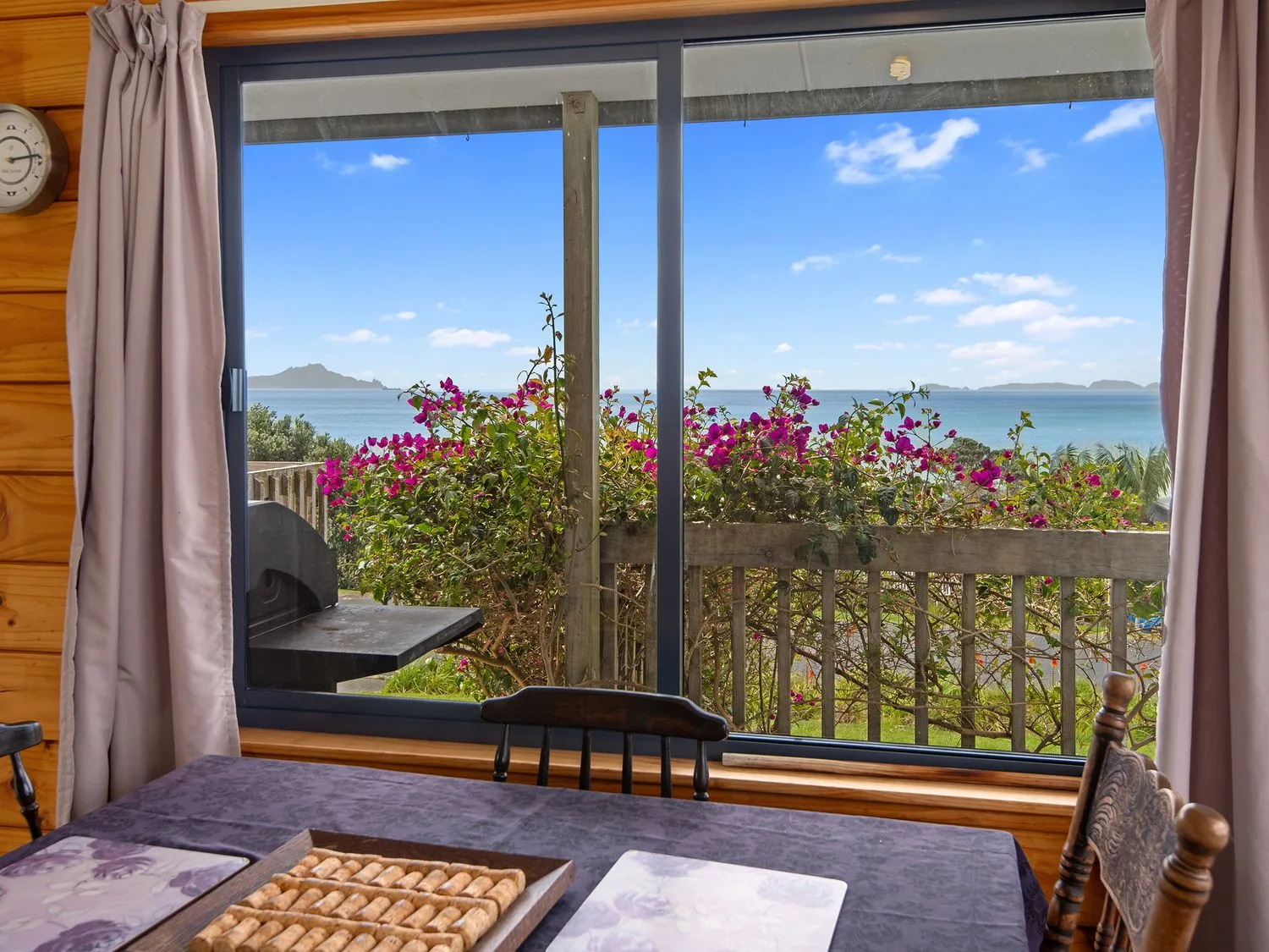 Un comedor con una mesa y sillas junto a una ventana con vistas a flores y al mar en Bay View Bach - Langs Beach Holiday Home, en Langs Beach