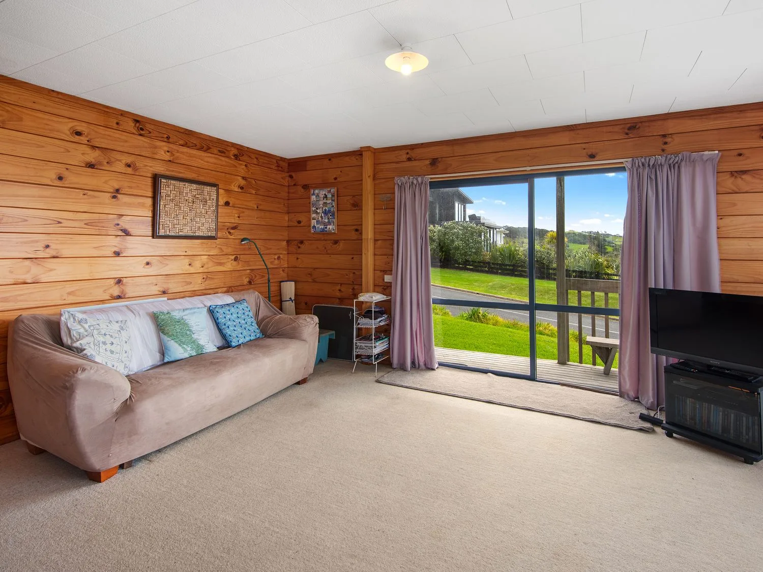 Una sala de estar con un sofá, cojines, una lámpara de pie, un televisor sobre un soporte y puertas correderas de cristal con cortinas en Bay View Bach Langs Beach