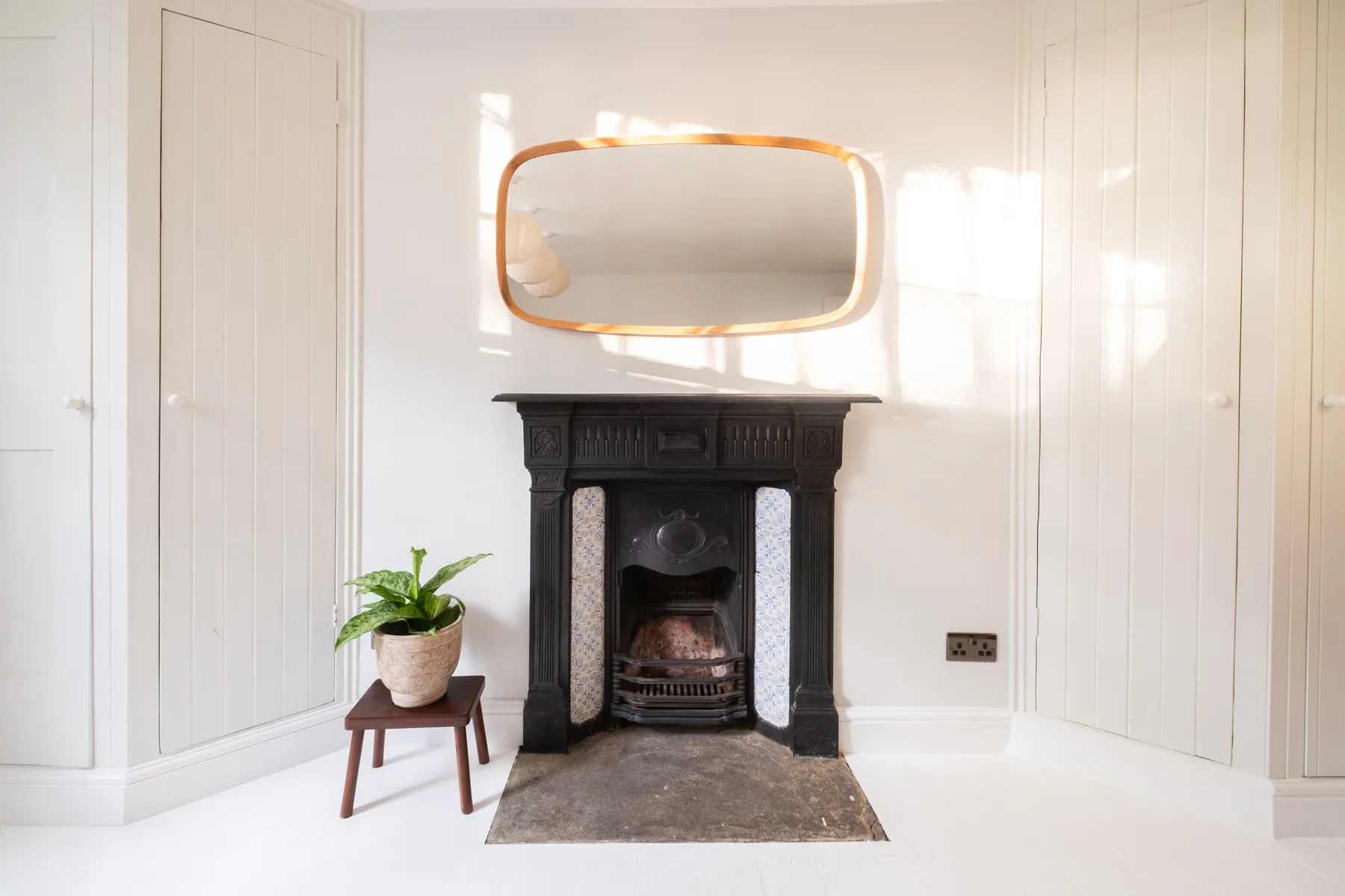 A bright room with a dark fireplace, wall mirror, and potted plant.