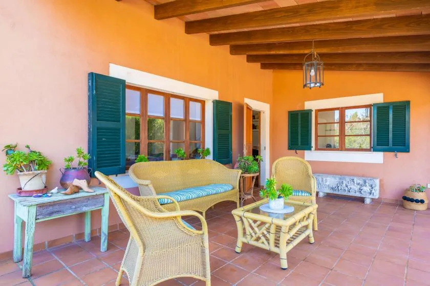A covered patio with wicker seating, potted plants, and windows with open shutters.