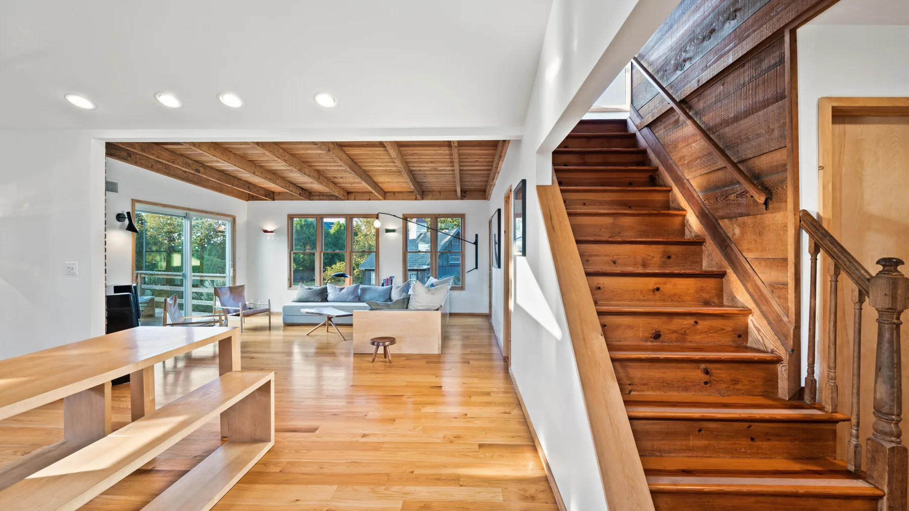Una luminosa sala de estar y comedor con grandes ventanas y una escalera de madera.