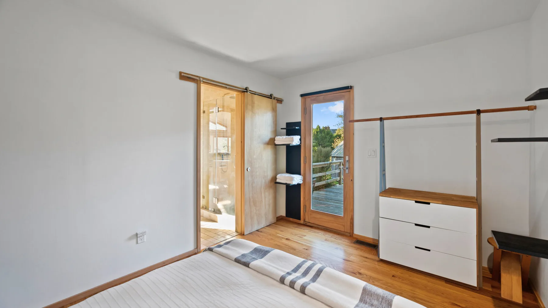 Un dormitorio luminoso con una cama, suelo de madera y una puerta acristalada al balcón.