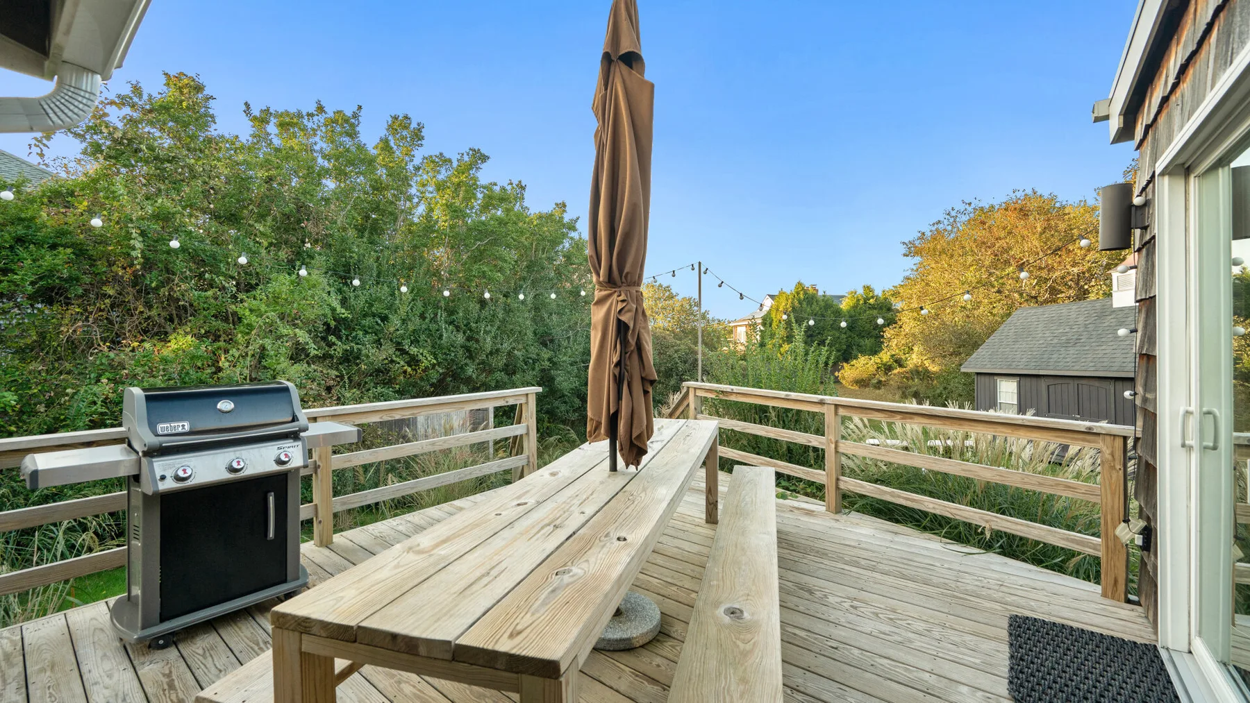 Una terraza exterior de madera con una parrilla, una mesa de picnic y vegetación alrededor.