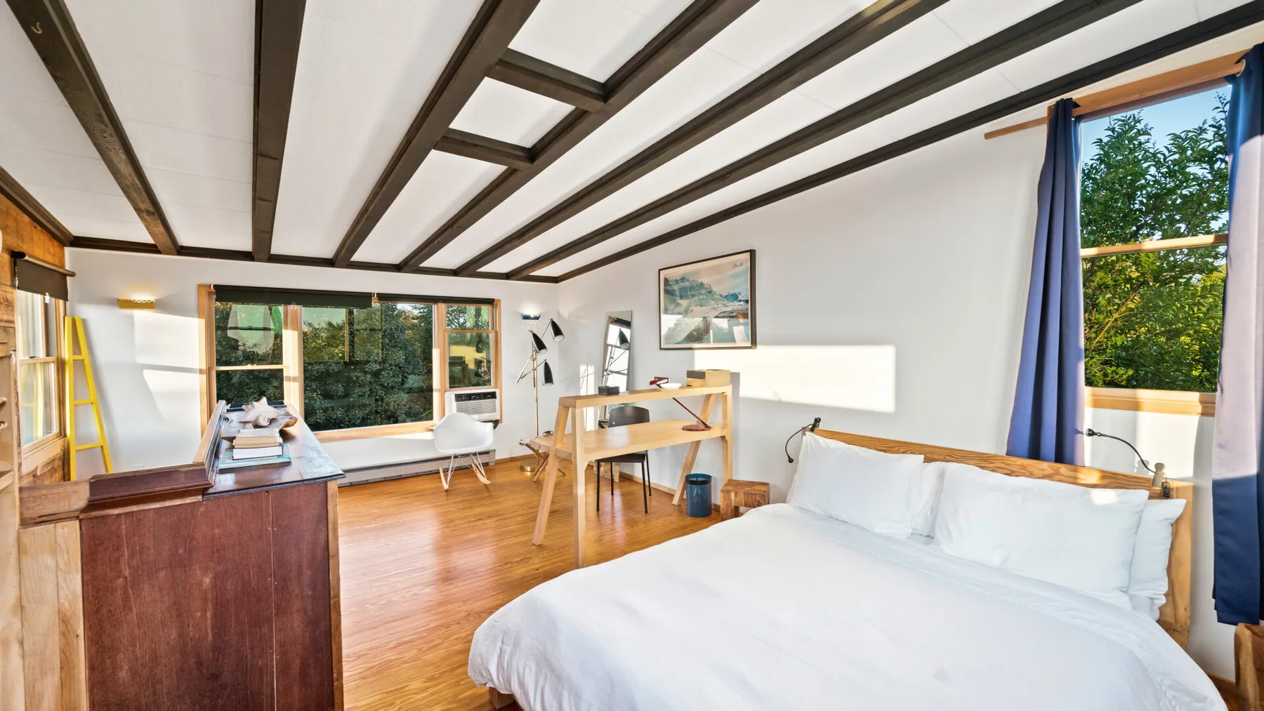 A bright bedroom with a large bed, a desk, and windows overlooking greenery.