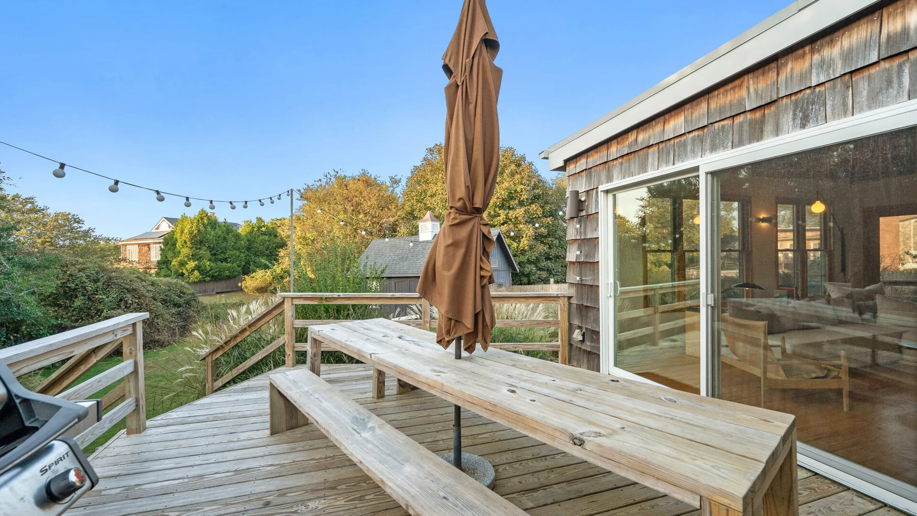 Una terraza de madera con una mesa larga de picnic y una sombrilla cerrada junto a la casa.