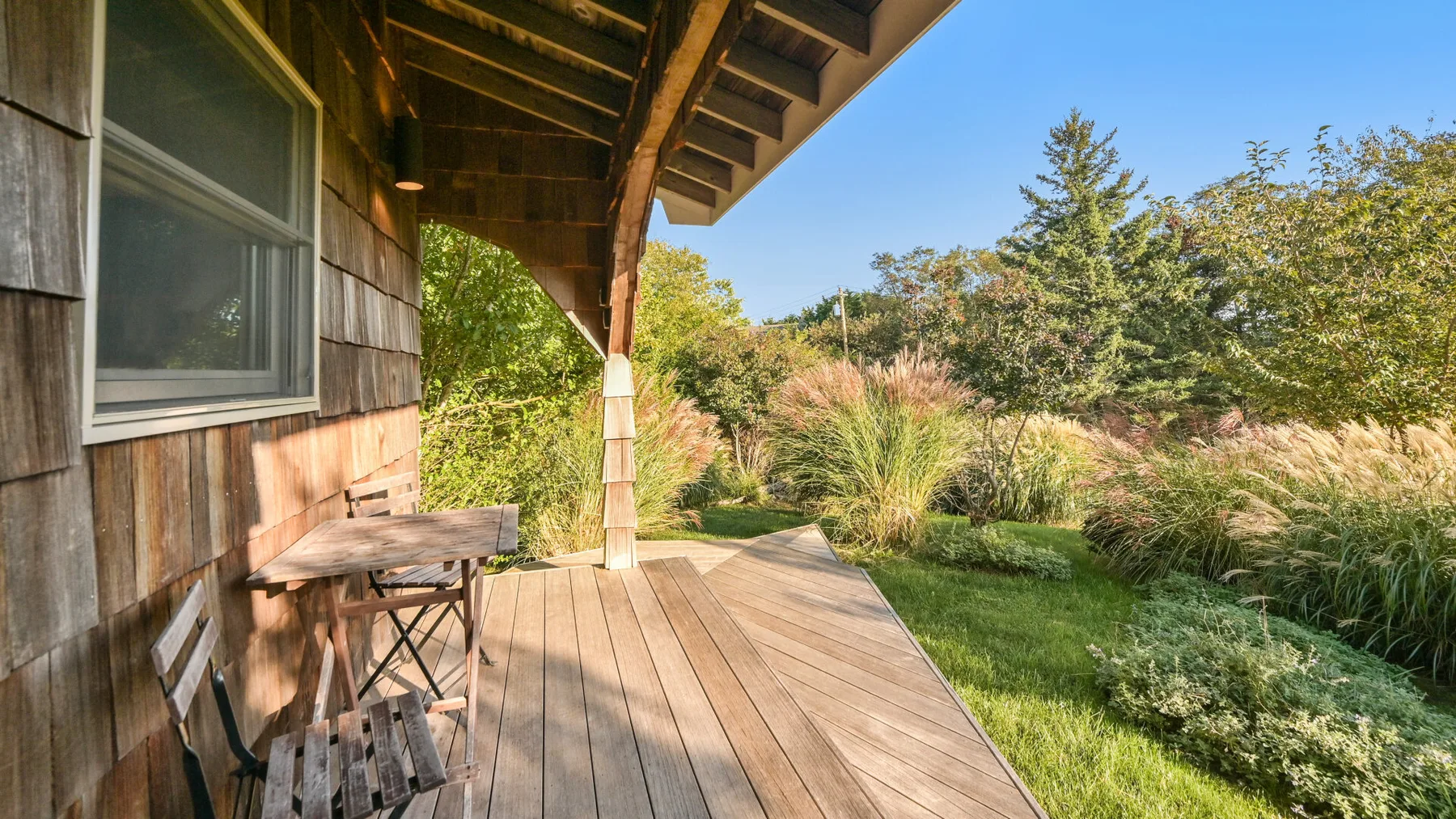 Una terraza de madera junto a una casa da a un jardín con hierbas y árboles.