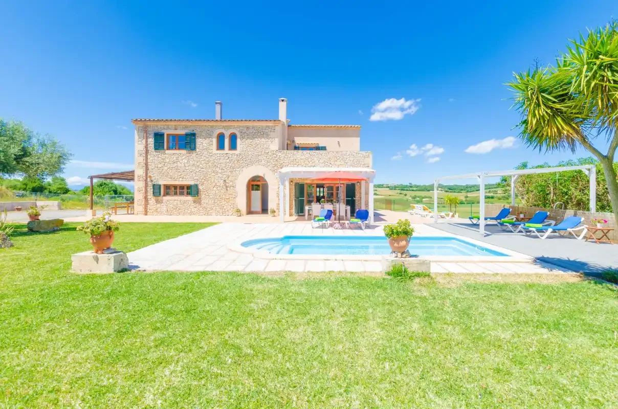 A stone house stands beside a swimming pool and lawn under a clear blue sky.