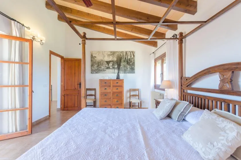 A bright bedroom with a large bed, wooden furniture, and exposed ceiling beams.