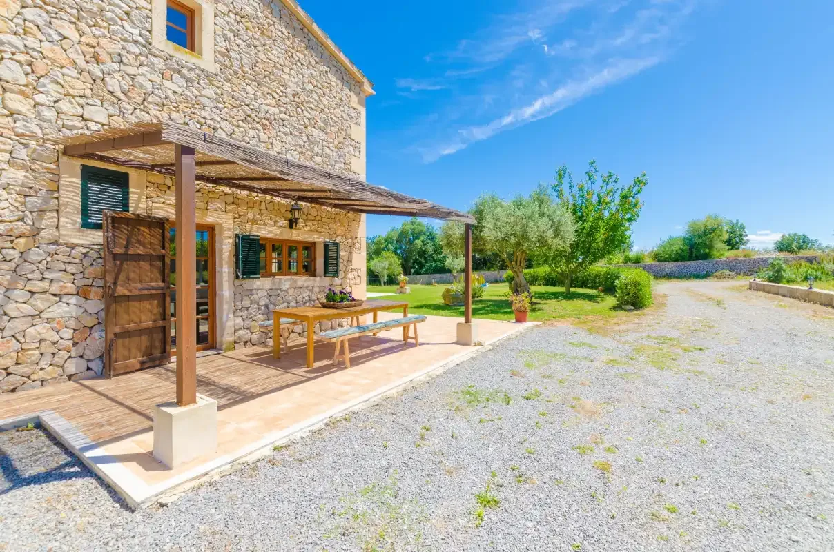 Stone house with a shaded patio and seating beside a gravel driveway.