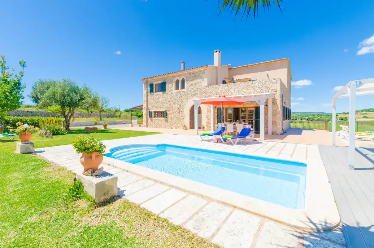 A stone house overlooks a swimming pool and patio in a sunny outdoor scene.