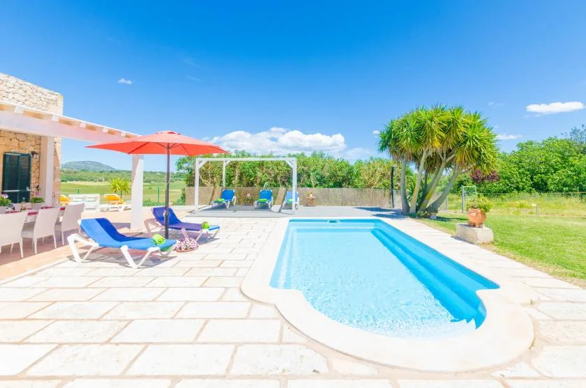 Outdoor swimming pool beside a paved patio with lounge seating and a red umbrella.