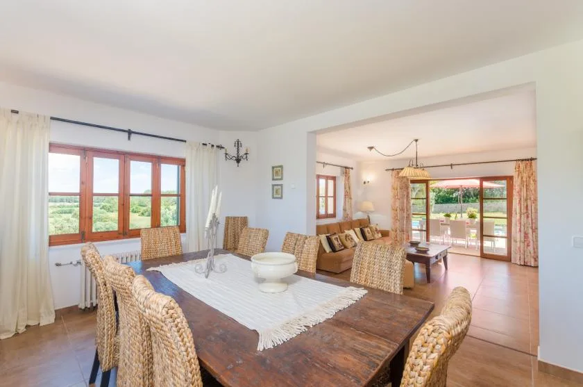 Dining room with a long wooden table, woven seating, and windows overlooking greenery.