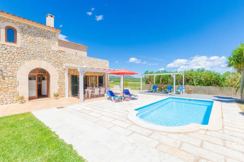 A swimming pool and paved patio sit beside a stone house under a clear blue sky.