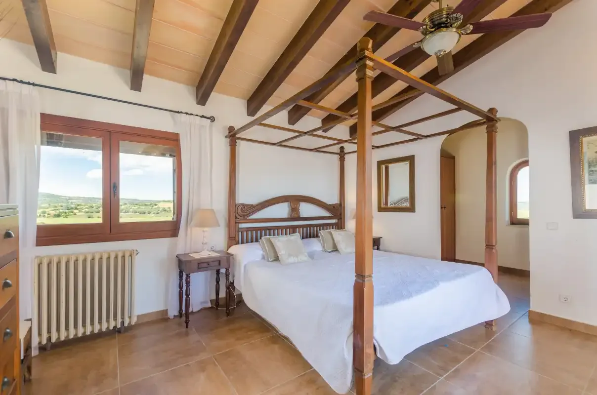 A bedroom with a four-poster bed, exposed ceiling beams, and a window overlooking the countryside.