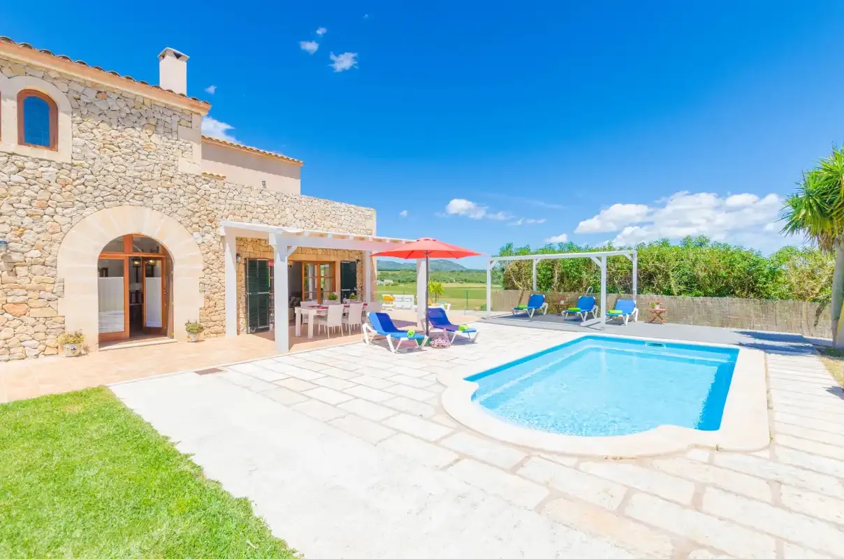 A stone house opens onto a sunny pool patio with loungers and an umbrella.