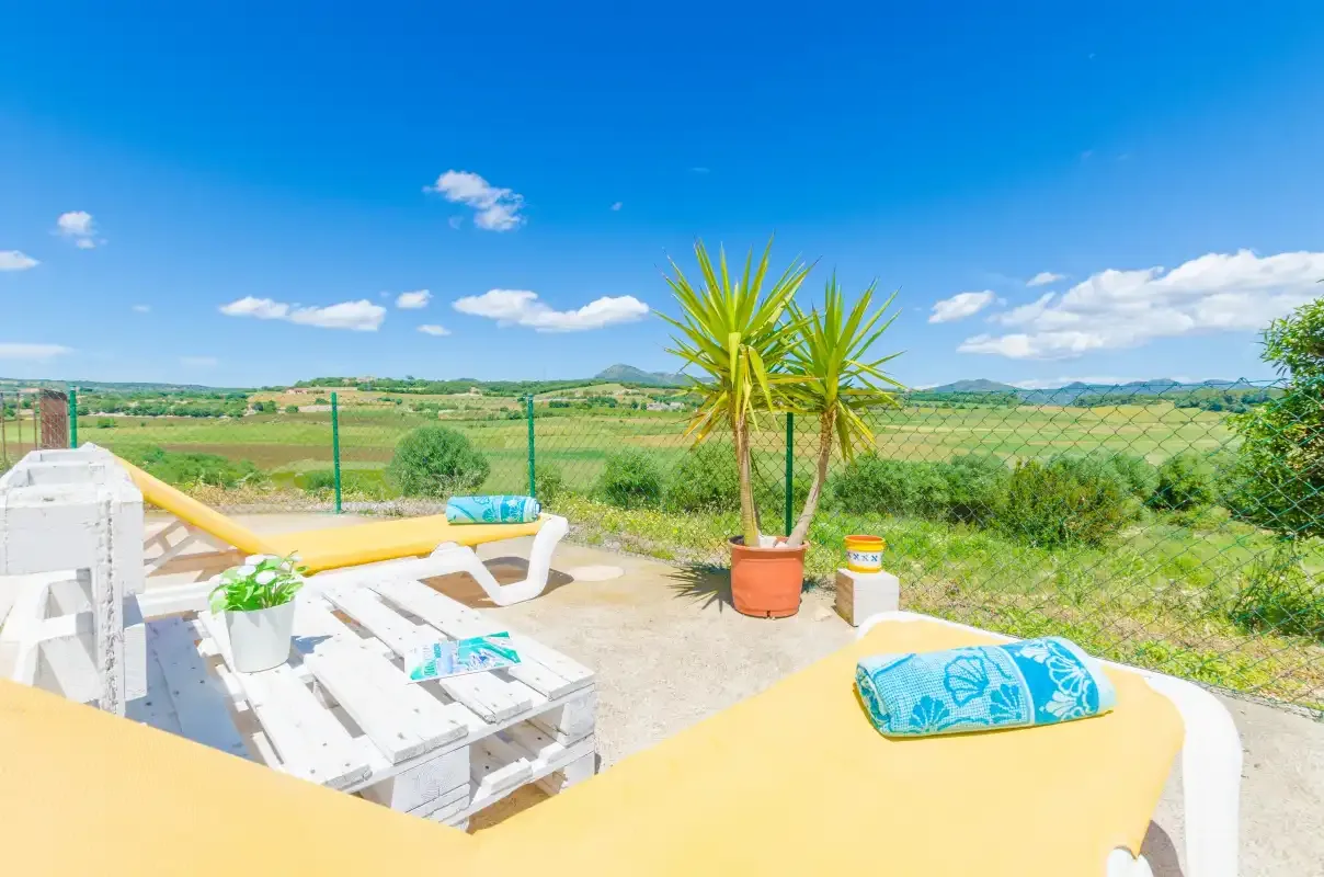 An outdoor patio has white seating, yellow cushions, and a potted plant overlooking fields.