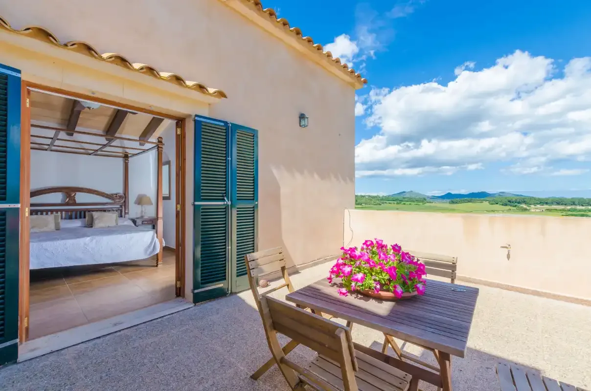 A sunny terrace with a wooden table and bright pink flowers beside an open bedroom.
