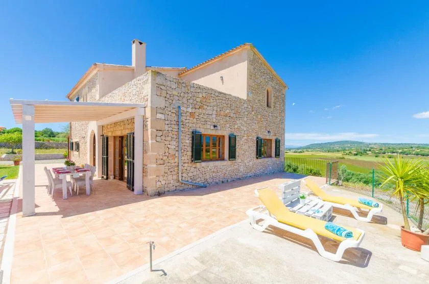 A stone house opens onto a sunny patio with outdoor seating and loungers.