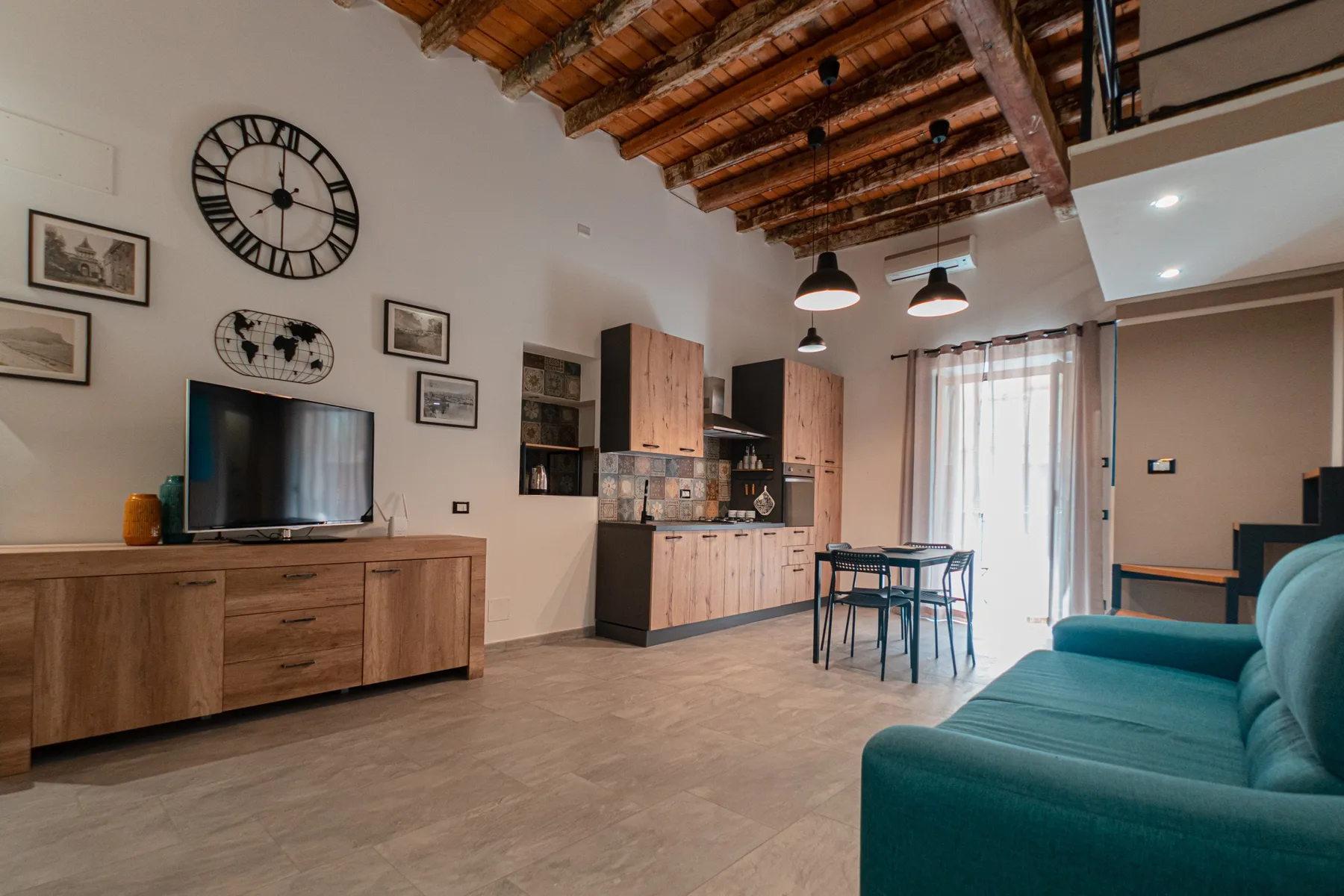 A living and dining area with a teal sofa, wall-mounted television, and compact kitchen.