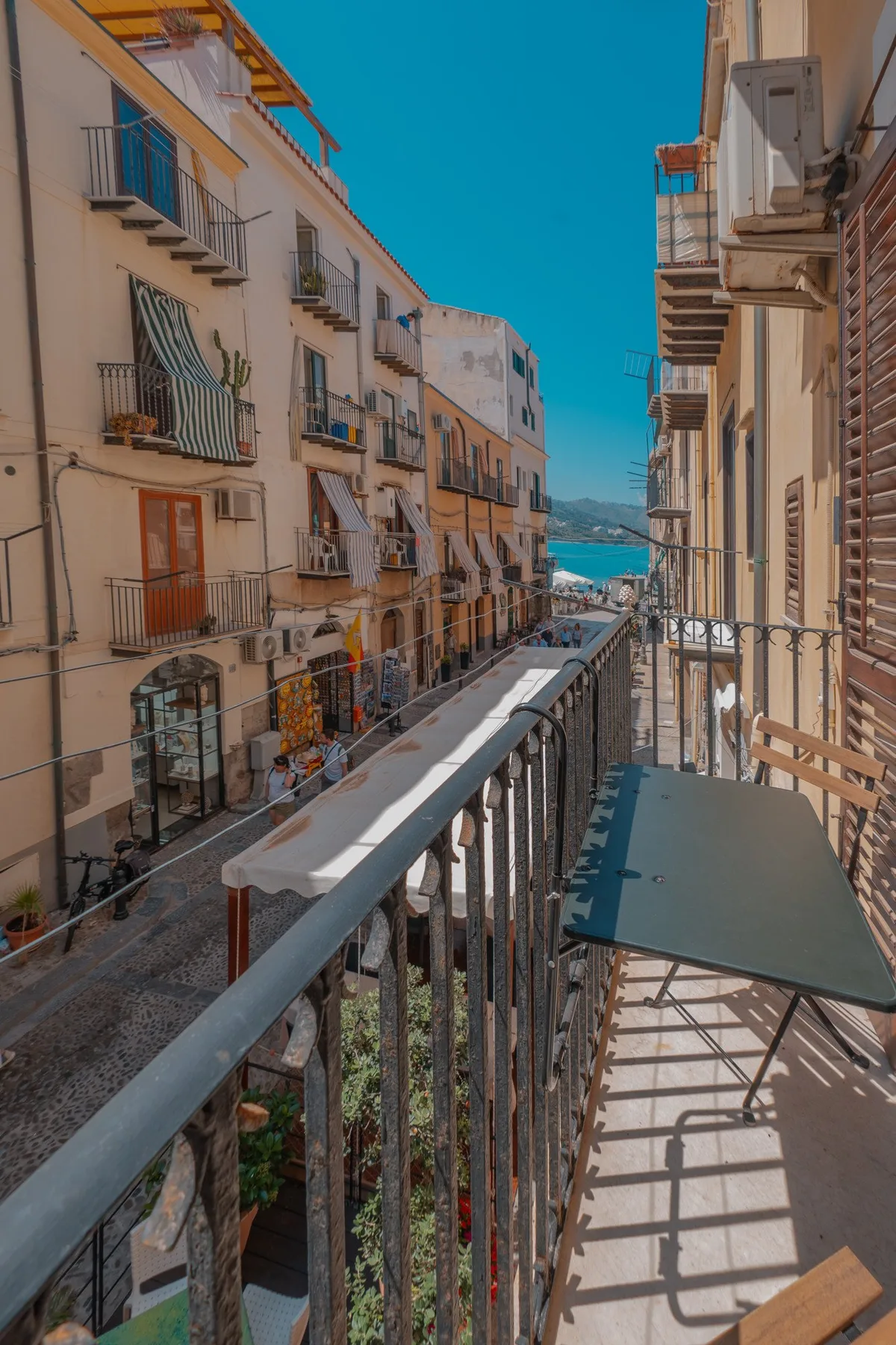 A narrow balcony overlooks a street lined with buildings and a glimpse of the sea.