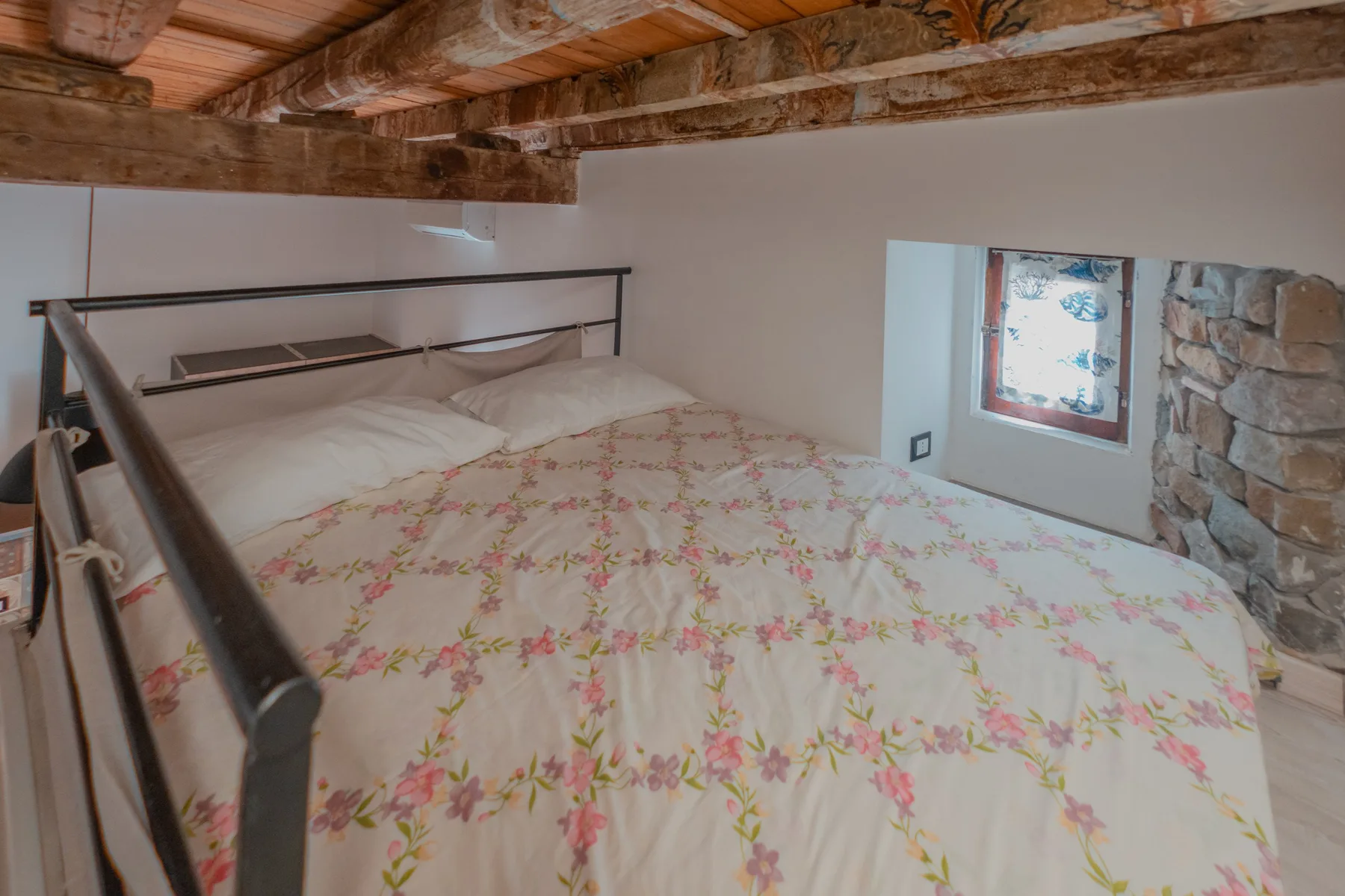 A bedroom with a floral bedspread beneath exposed wooden ceiling beams.