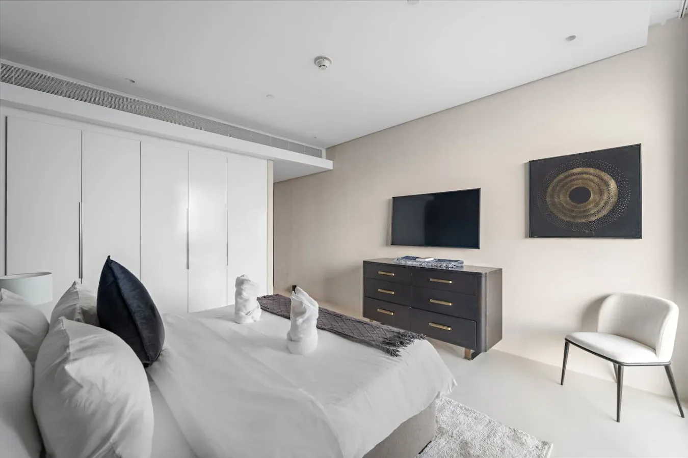 Un dormitorio con una cama tendida, una cómoda oscura y un televisor en la pared.