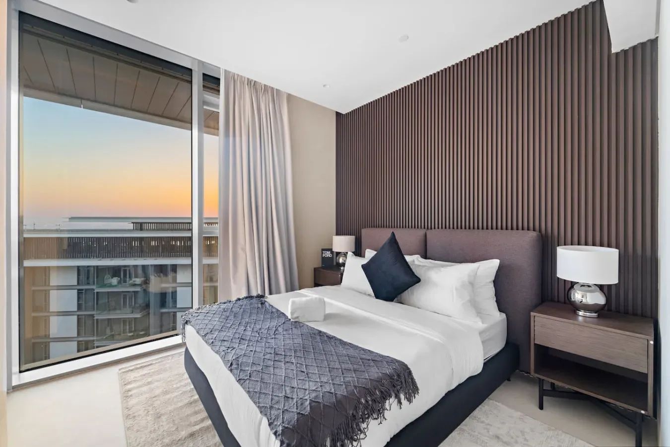 Un dormitorio con una cama bien tendida junto a una ventana grande al atardecer.