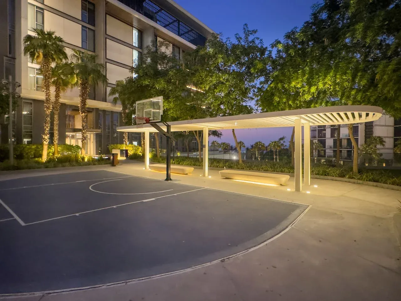 Una cancha de baloncesto al aire libre con una canasta junto a una zona cubierta para sentarse al anochecer.