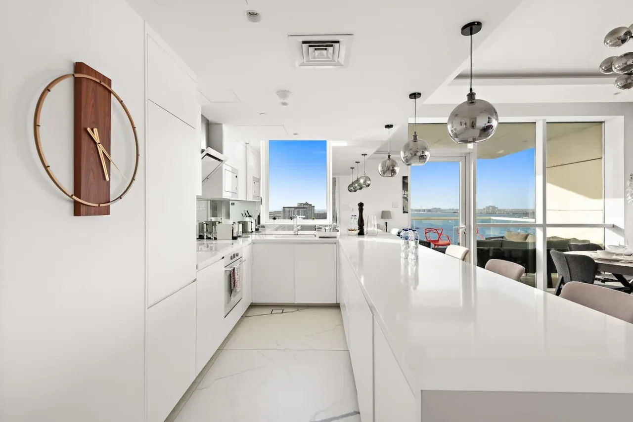 Una cocina blanca y luminosa con una isla larga y lámparas colgantes.