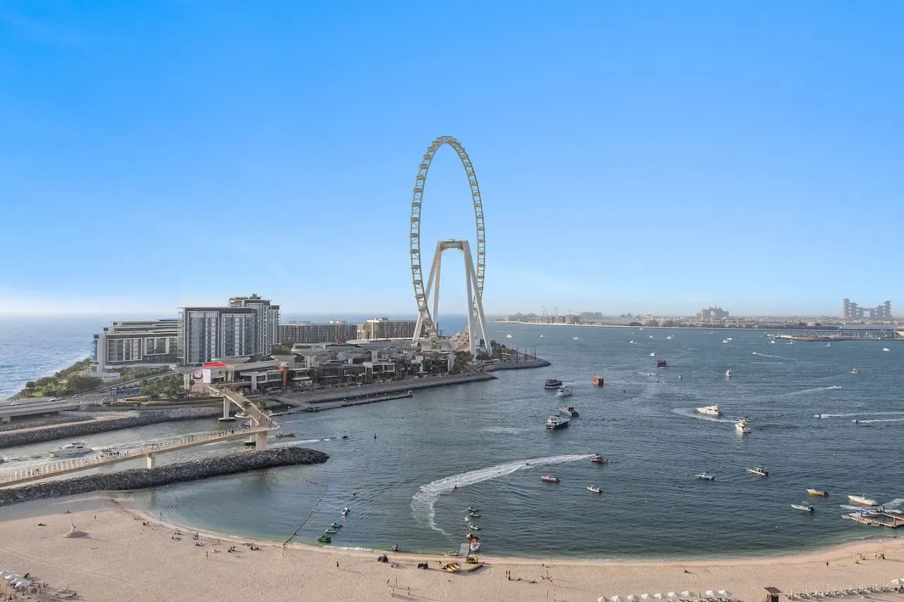 Un paseo costero con una gran noria, edificios y barcos sobre el agua.