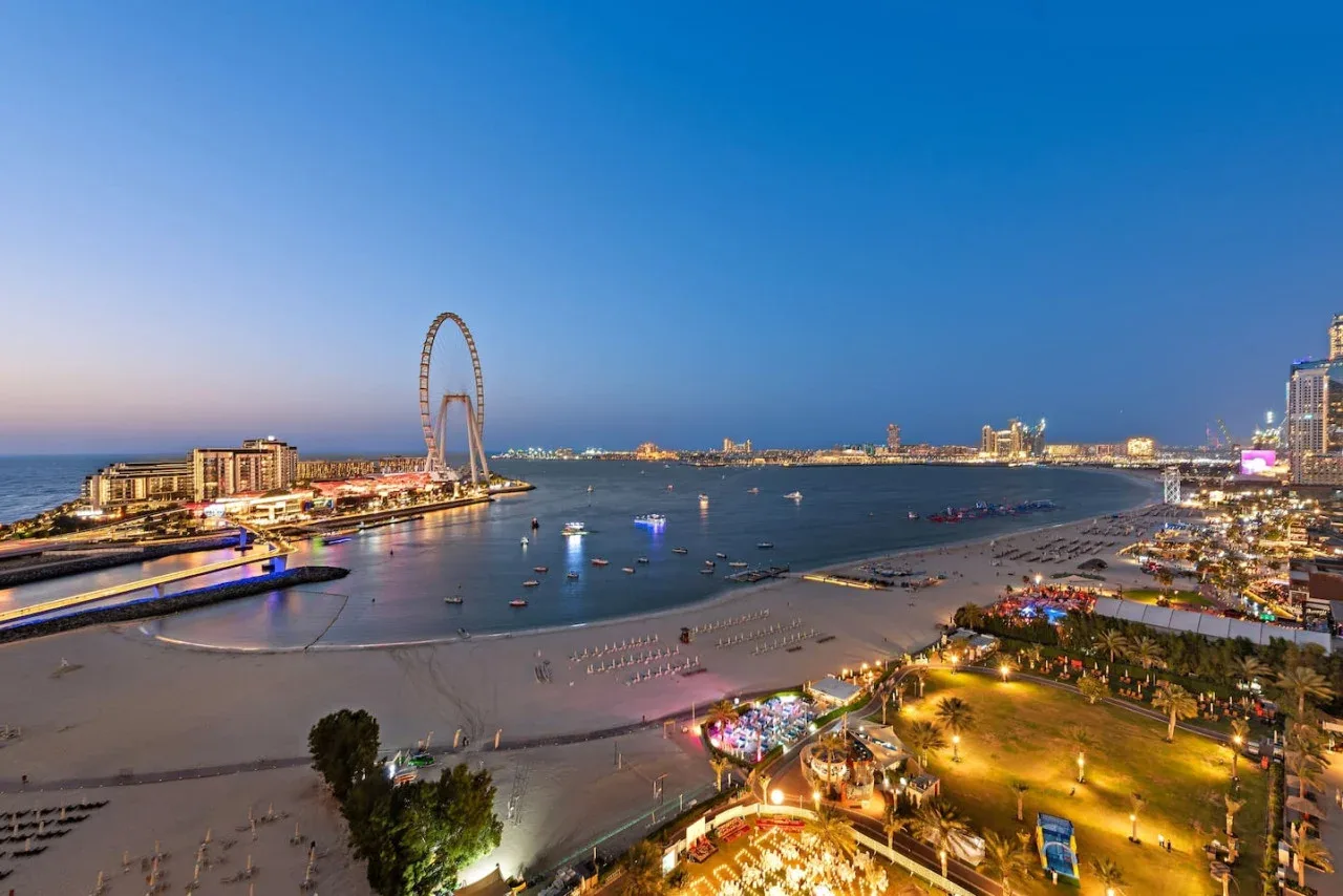 Un paseo marítimo al anochecer, con una playa de arena, barcos y edificios iluminados.