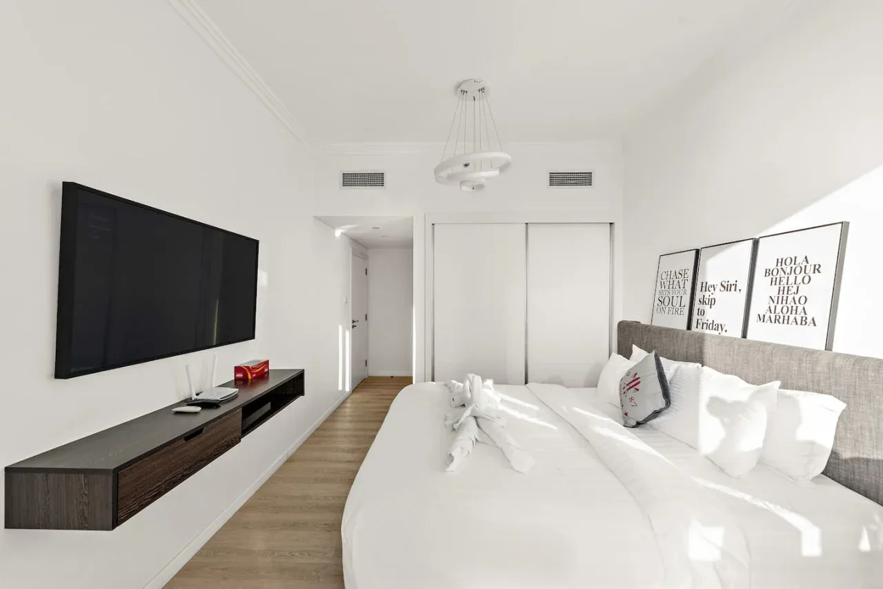 Un dormitorio luminoso con una cama grande, un televisor en la pared y láminas enmarcadas.