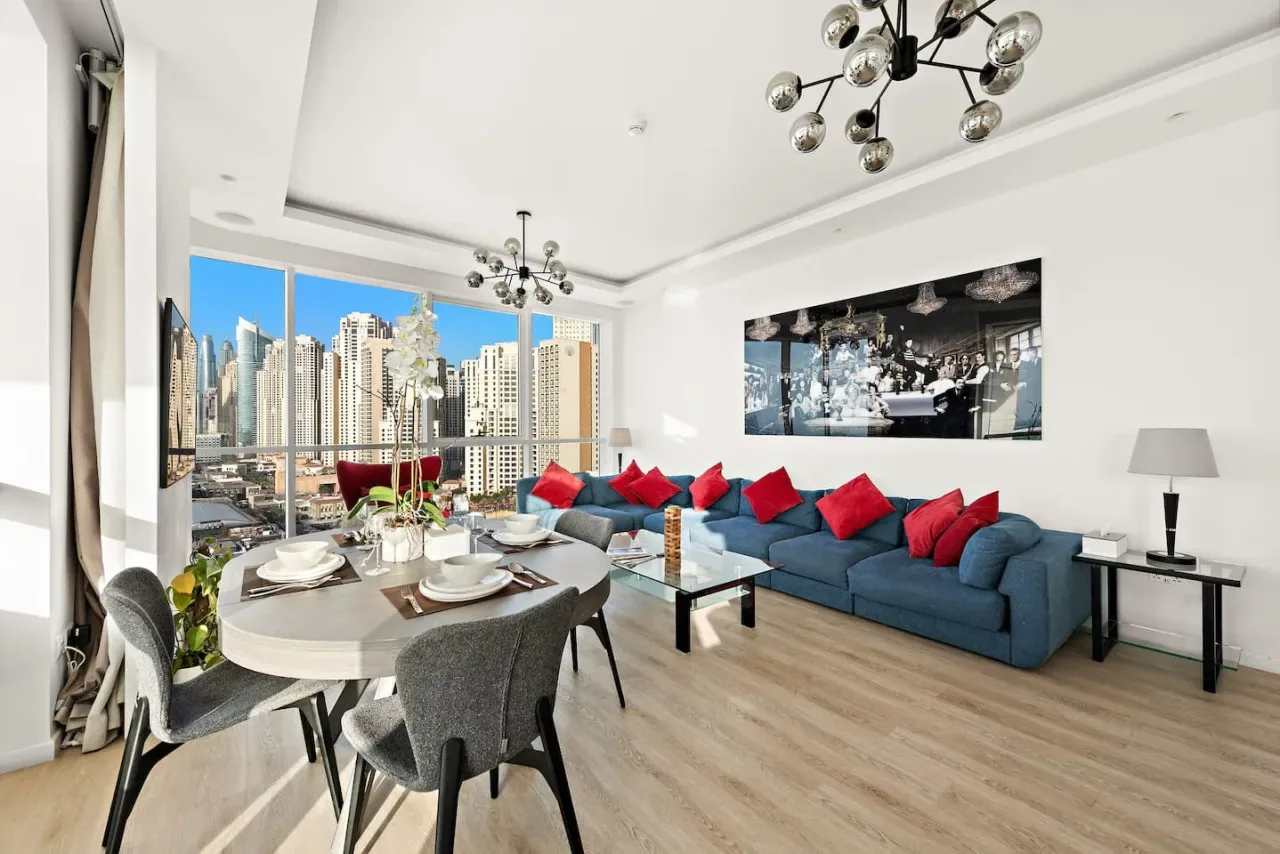Una luminosa sala de estar y comedor con asientos azules y grandes ventanas con vistas a edificios de la ciudad.