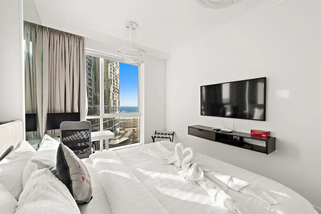 Un dormitorio luminoso con una cama frente a un televisor en la pared y una ventana grande.