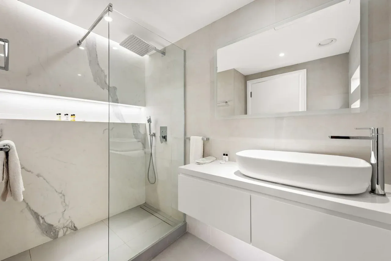 Un baño luminoso con una ducha de vidrio y un lavabo blanco sobre la encimera.