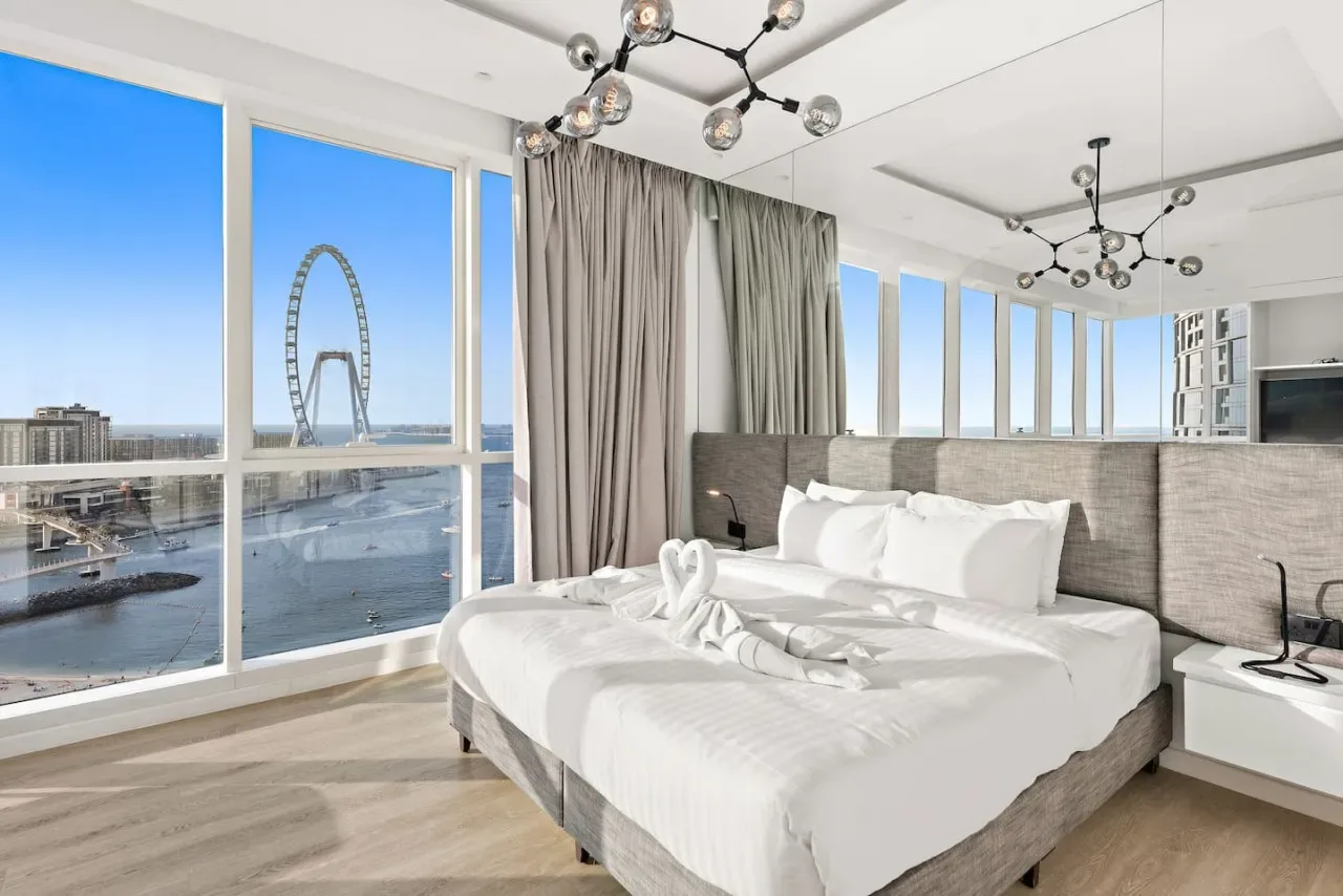 Un dormitorio luminoso con ropa de cama blanca, cabecero gris y grandes ventanales.