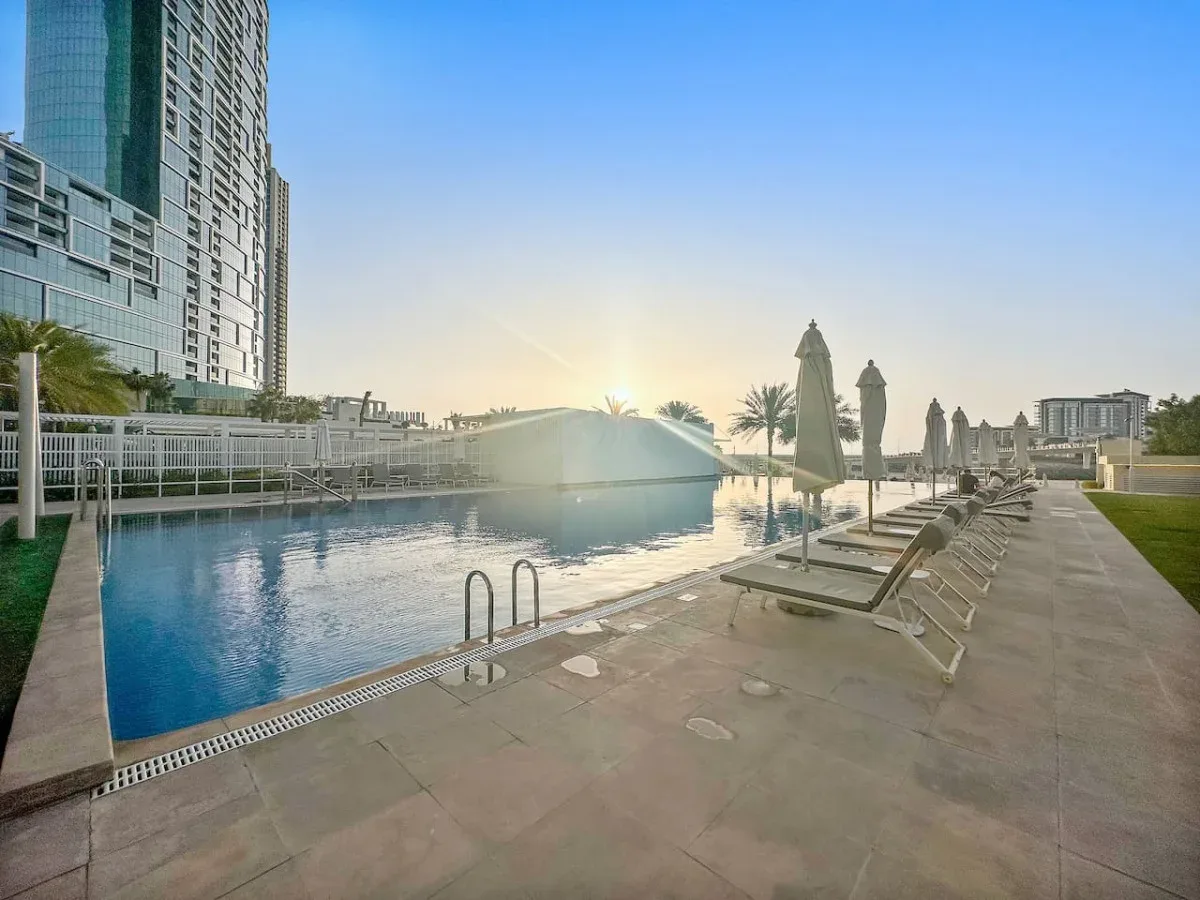 Una piscina al aire libre junto a una terraza pavimentada con tumbonas y sombrillas cerradas.