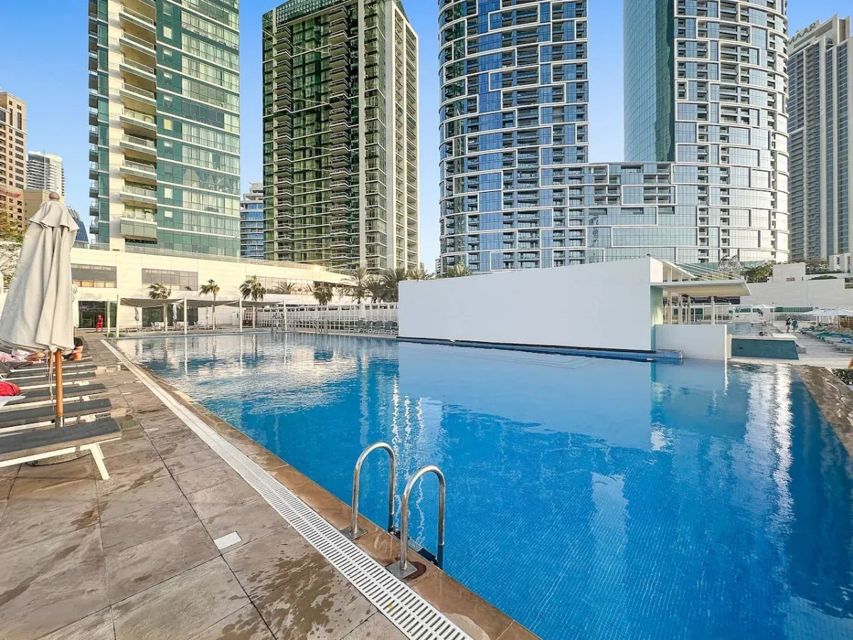 Una piscina al aire libre está junto a una terraza pavimentada, con edificios altos al fondo.
