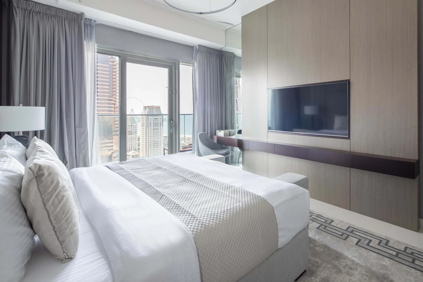 Un dormitorio con una cama bien tendida junto a grandes ventanas con edificios cercanos a la vista.