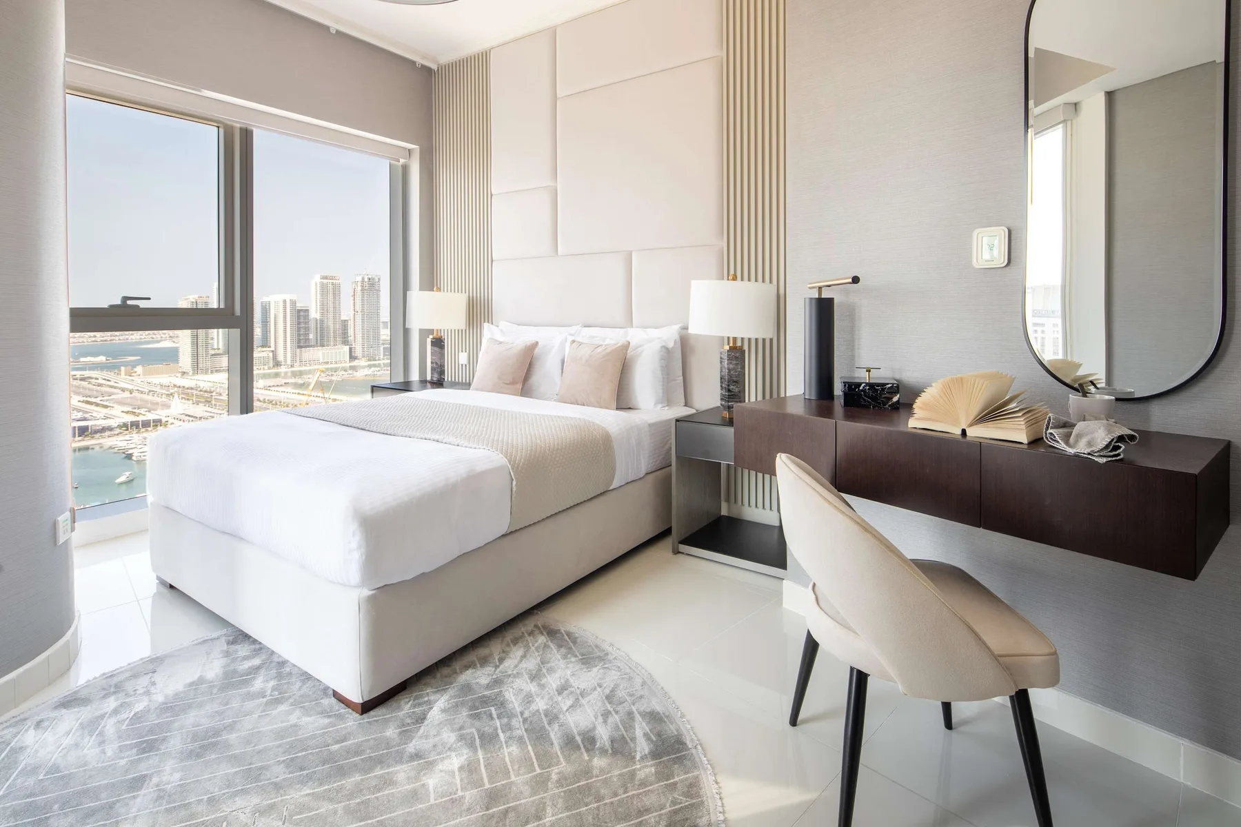 Un dormitorio luminoso con una cama bien tendida junto a grandes ventanas con vistas a la ciudad.